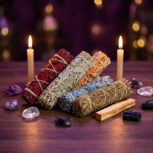 Professional product photography, square 1:1. Sacred spiritual shop display on a dark wood table: five smudge sage bundles arranged beautifully together — one deep red dragon's blood sage, one classic green-gray white sage, one warm orange pumpkin-toned sage, one soft blue sage, and one cedar bundle with brownish-green needles — plus a small piece of palo santo wood beside them. Around the bundles: two lit chime candles, a few tumbled crystals (amethyst, clear quartz, black tourmaline), soft mystical purple and gold ambient light, shallow depth of field, rich dark purple background bokeh, elegant Sacred Ground metaphysical store aesthetic, high detail, no text, no watermark, no logos.